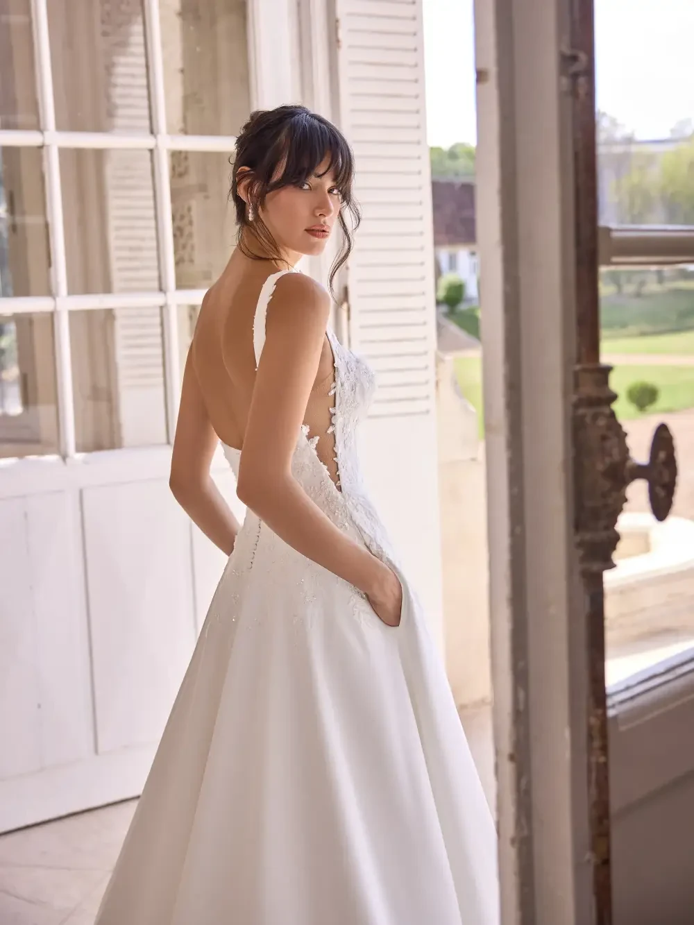 A woman in a wedding dress stands with her back facing the camera, featuring a low back and intricate lace detailing, with pockets in the skirt. Natural light streams through large windows, casting a soft glow in the room.