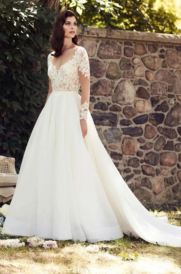 Model is wearing a romantic long sleeve A-line gown with an Alencon lace bodice, open back, and flowing organza skirt.