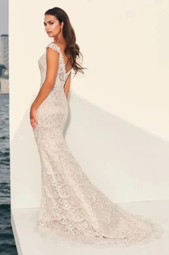 Model is wearing a fit and flare lace gown with a V-neckline, off-the-shoulder cap sleeves, champagne satin lining, and a delicate belt.