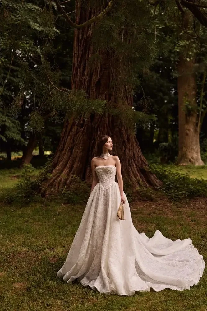 A woman in a strapless white wedding dress stands gracefully in front of a large tree in a lush green setting.