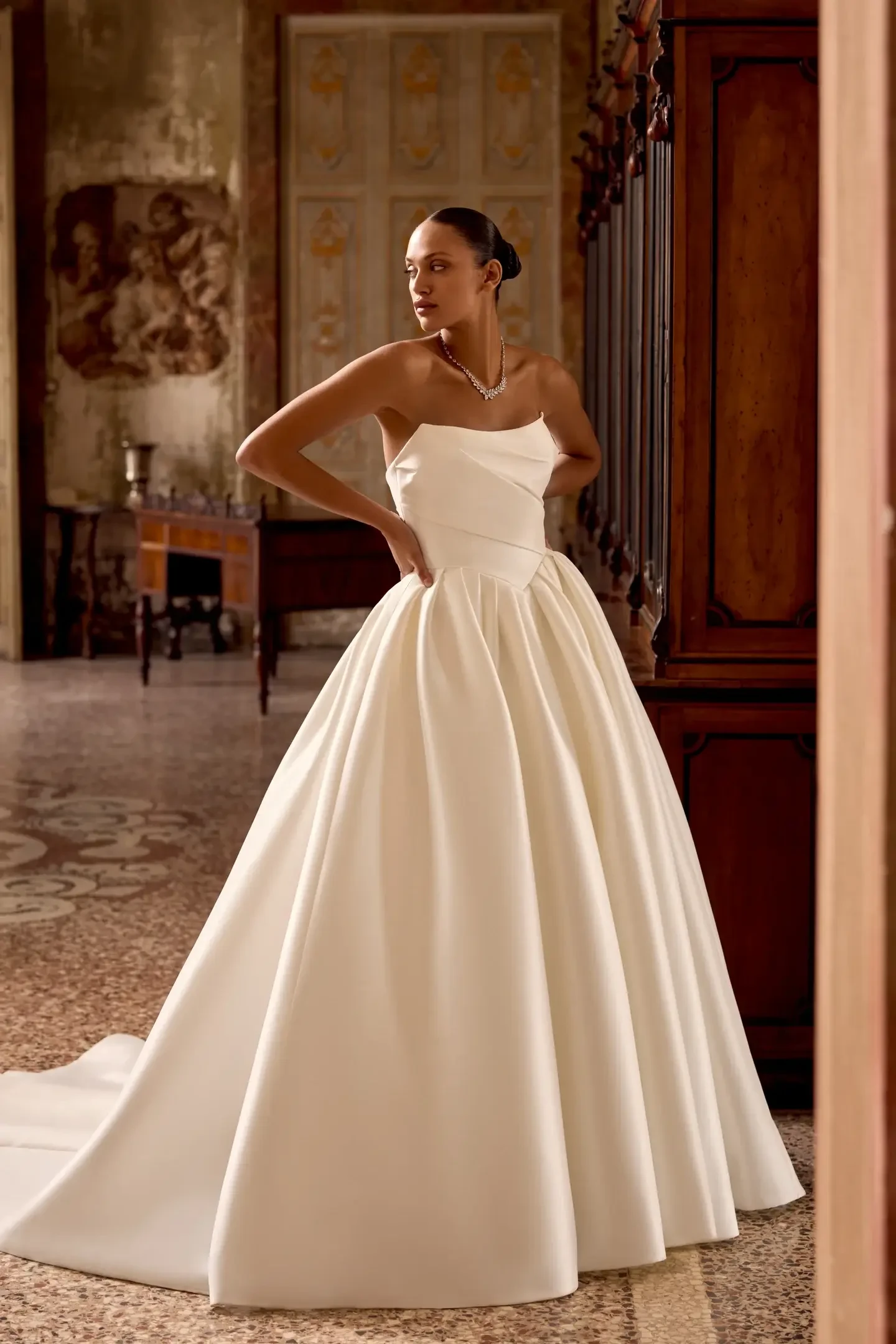 A woman stands elegantly in a luxurious room, wearing a strapless white ball gown with a full skirt. The ambiance is regal and sophisticated.