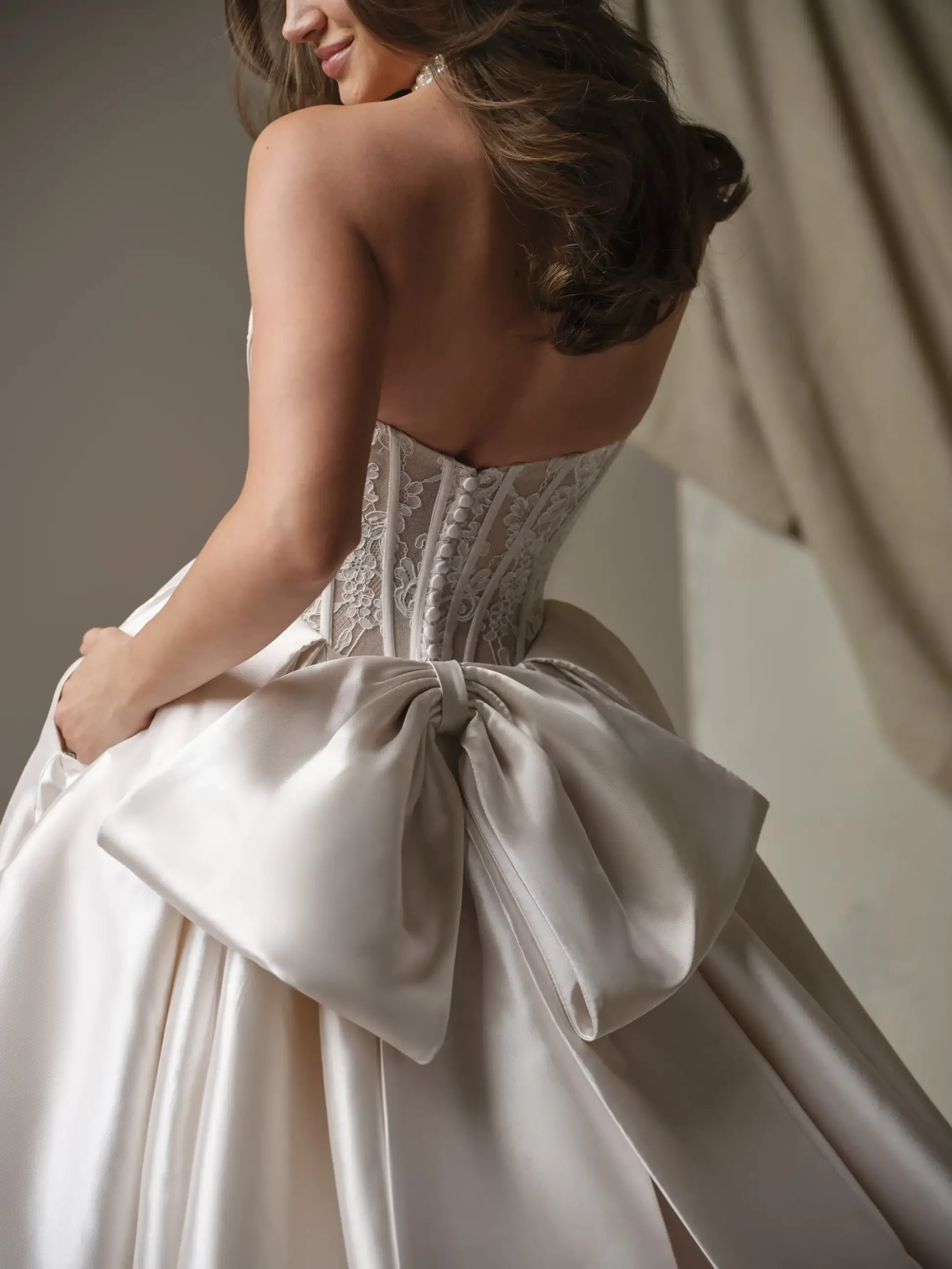 Back view of a woman in an elegant ivory gown featuring a lace bodice and a large satin bow. Her wavy hair cascades down, conveying a romantic tone.