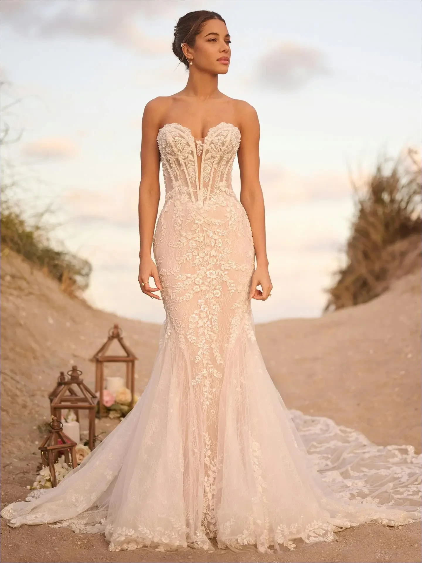 A woman wearing a stunning strapless wedding gown with intricate lace details, standing on a sandy path surrounded by greenery.