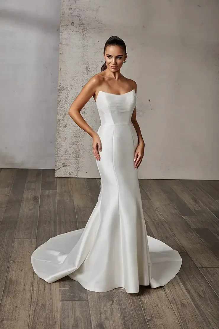 A woman in a sleek, strapless white wedding gown stands on a wooden floor. The dress has a smooth texture and long train, set against a textured gray wall.