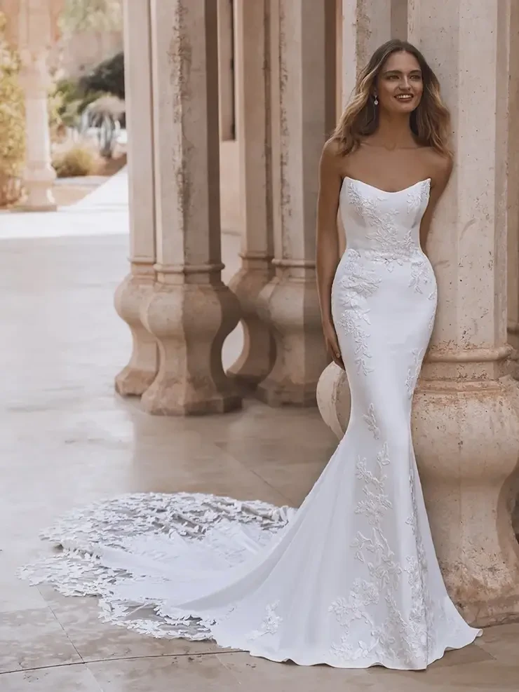 A woman in an elegant, strapless white wedding dress with lace details and a long train stands smiling by stone columns in a sunlit, serene courtyard.