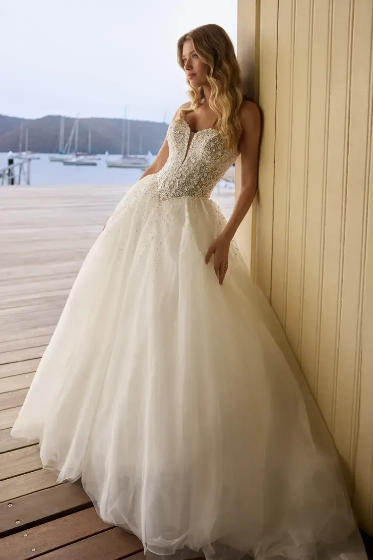 A woman in a strapless, embellished wedding dress stands against a wooden wall, with a scenic waterfront in the background.