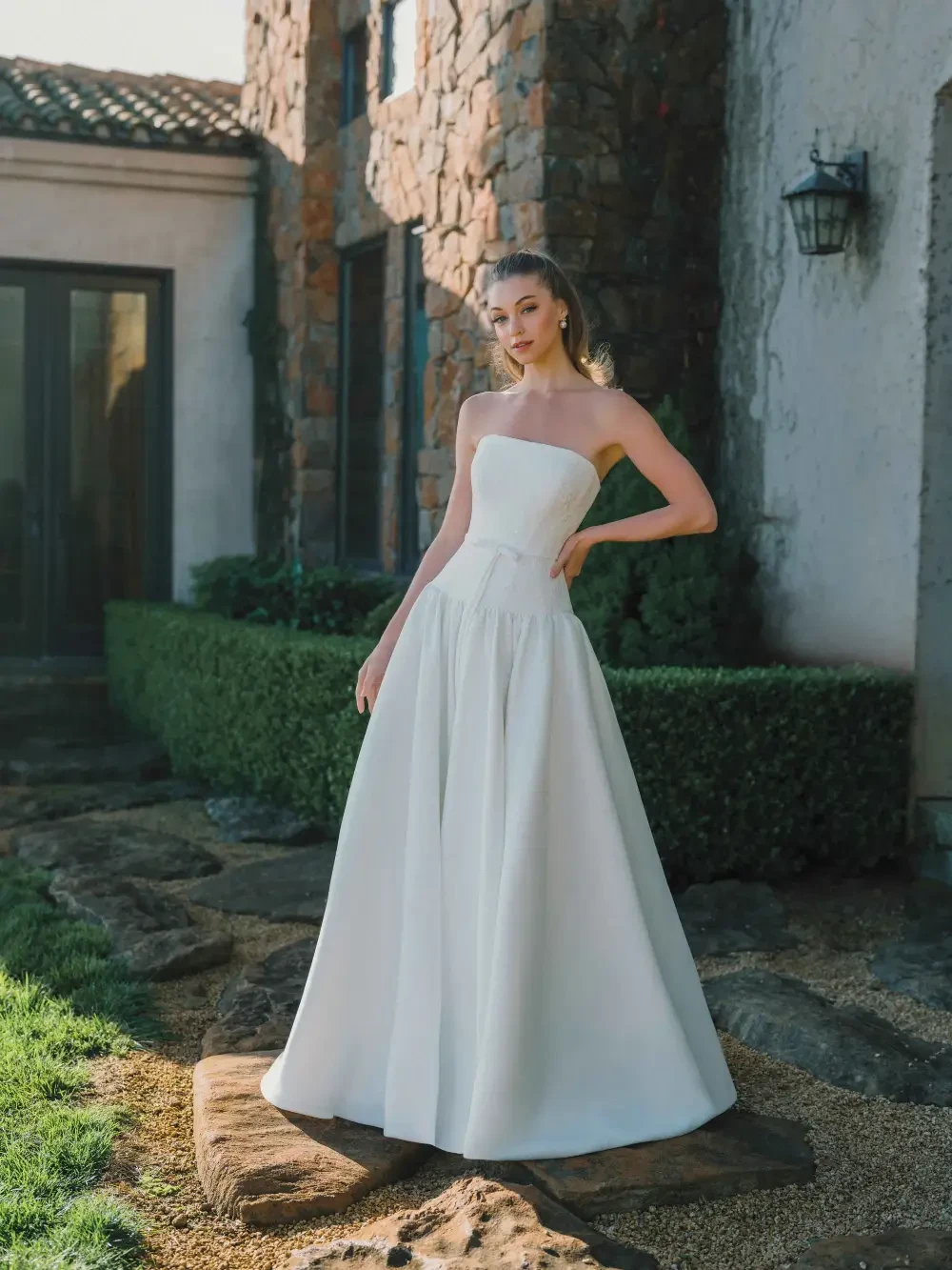 A woman in a strapless white wedding gown stands gracefully outside a stone building, with lush greenery and sunlight enhancing the scene.