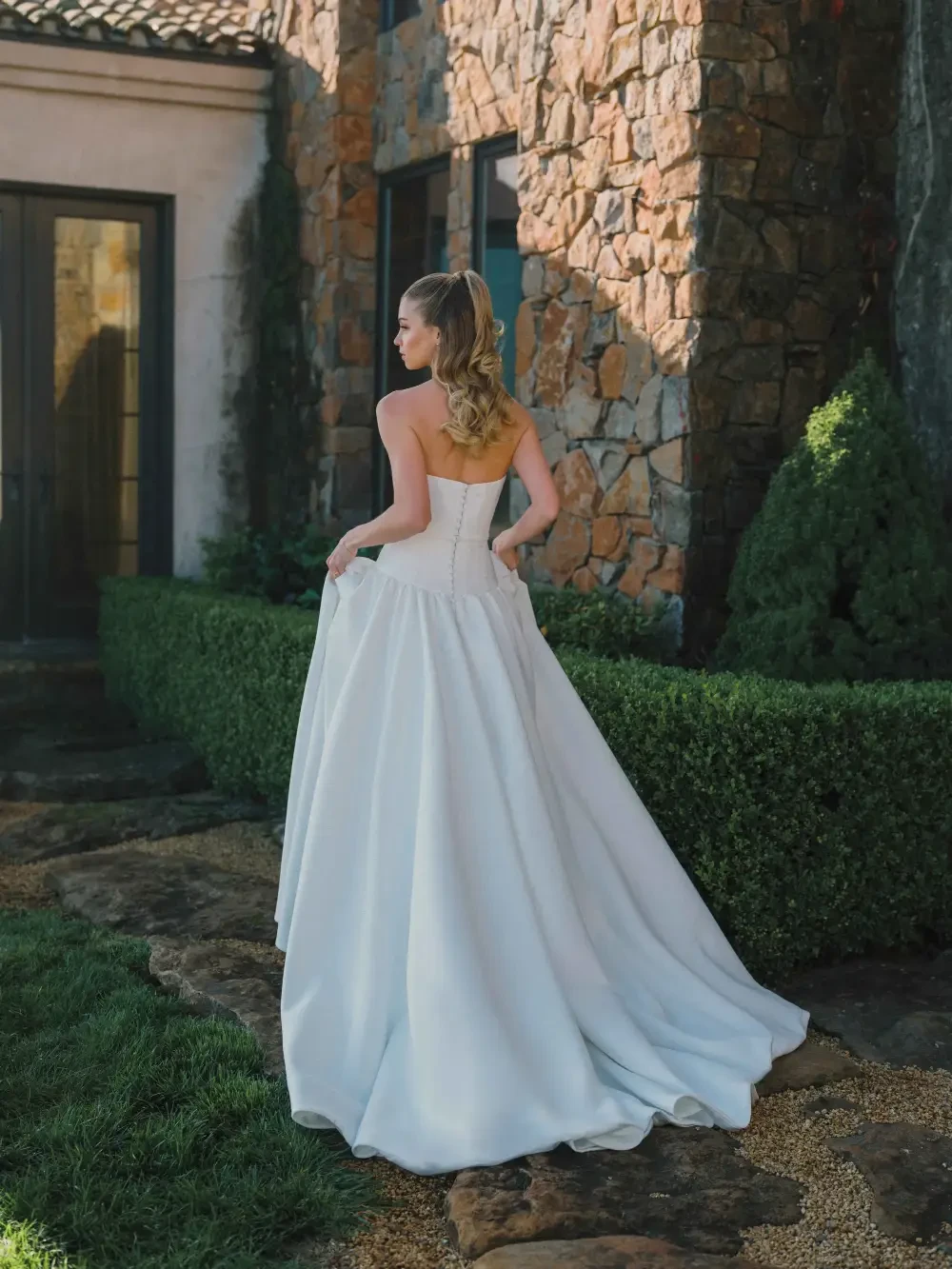 A woman in a strapless white wedding gown stands gracefully outside a stone building, with lush greenery and sunlight enhancing the scene.