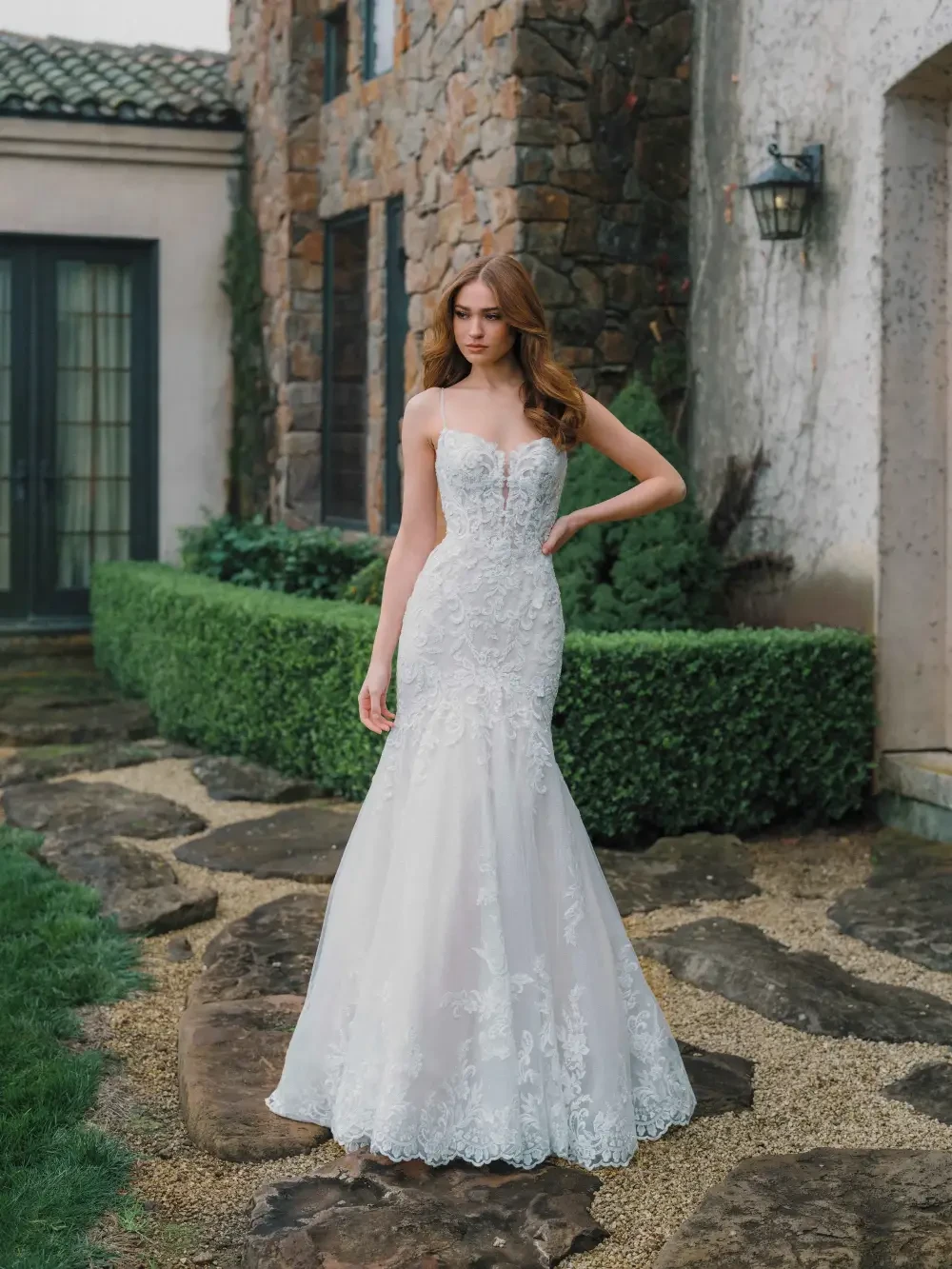 A woman in a strapless wedding gown stands on a stone pathway in a garden, with stone buildings in the background.