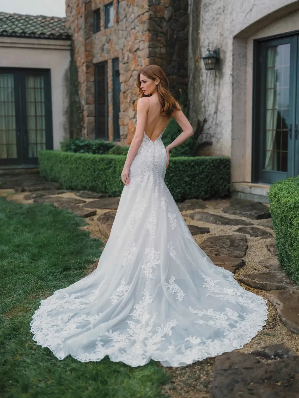 A woman in a strapless wedding gown stands on a stone pathway in a garden, with stone buildings in the background.