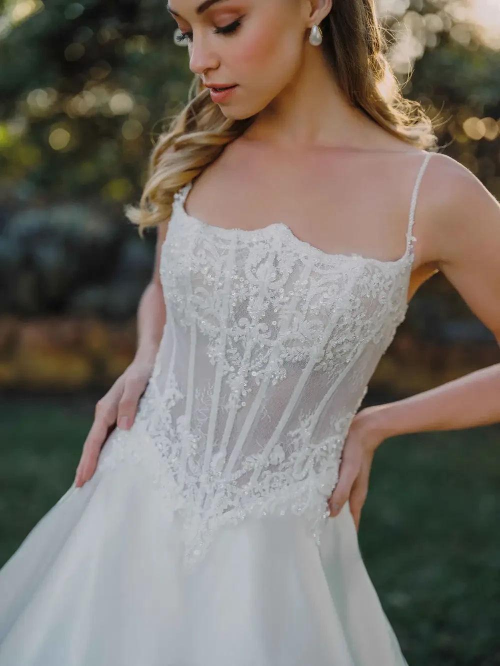 A woman wearing an elegant wedding dress with intricate lace details, standing outdoors with a serene expression.
