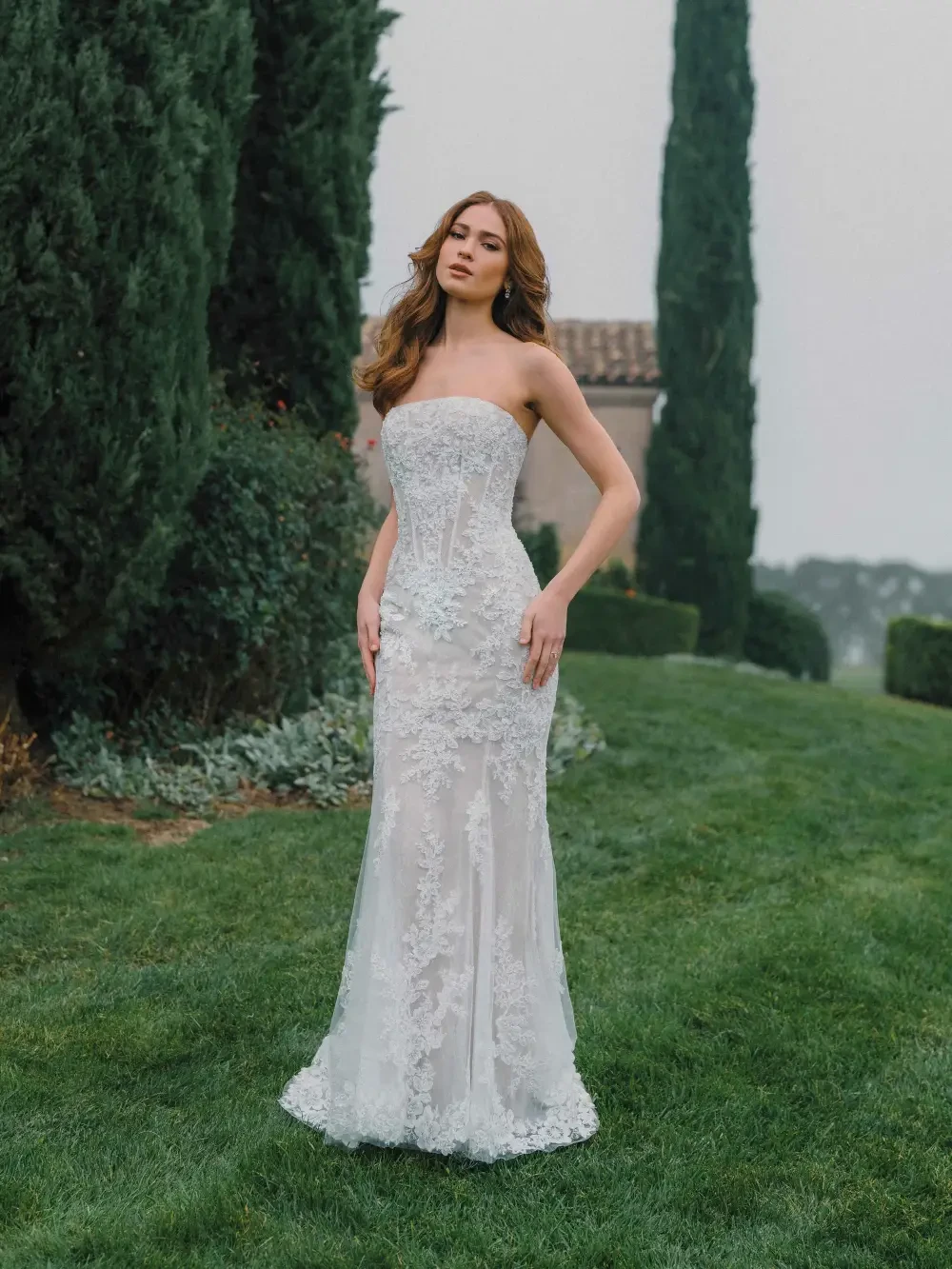 A woman in a strapless, elegant wedding gown stands on a lush green lawn surrounded by tall trees and hedges.
