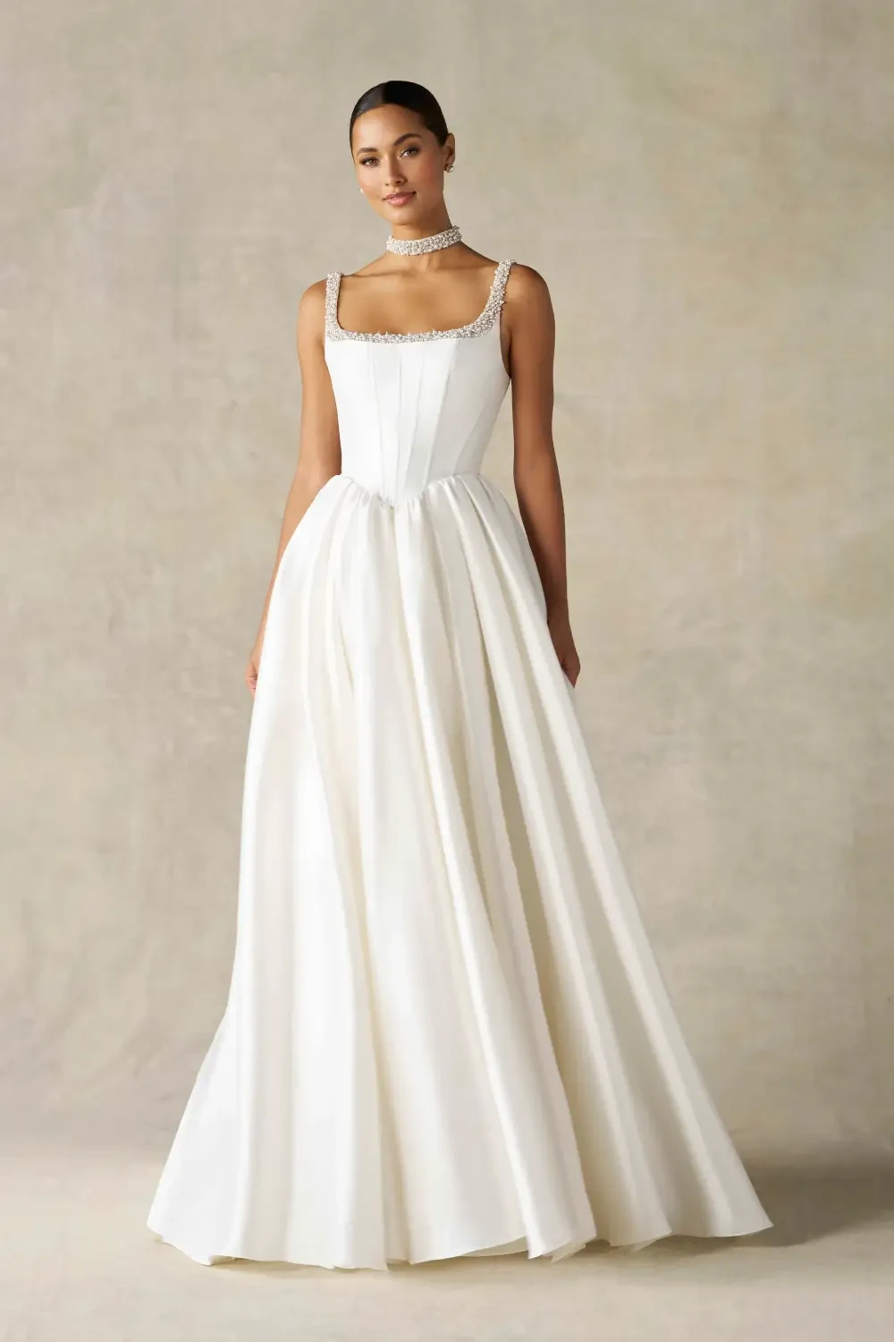 A model wearing a beautiful white gown with a fitted bodice, square neckline, and a flowy skirt, complemented by a pearl choker necklace, posed against a textured neutral background.