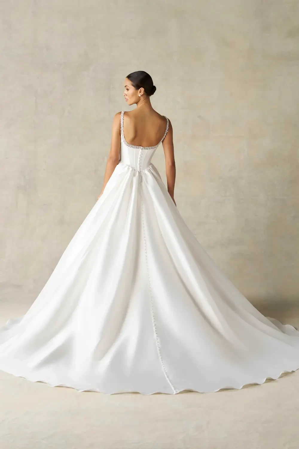 A model wearing a beautiful white gown with a fitted bodice, square neckline, and a flowy skirt, complemented by a pearl choker necklace, posed against a textured neutral background.