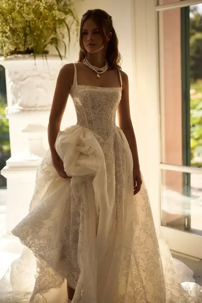 A model wearing an elegant white wedding dress standing indoors, with soft natural light illuminating the scene.