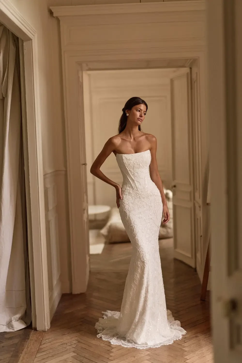A model in a strapless, fitted white wedding gown standing in a softly lit room with elegant decor.