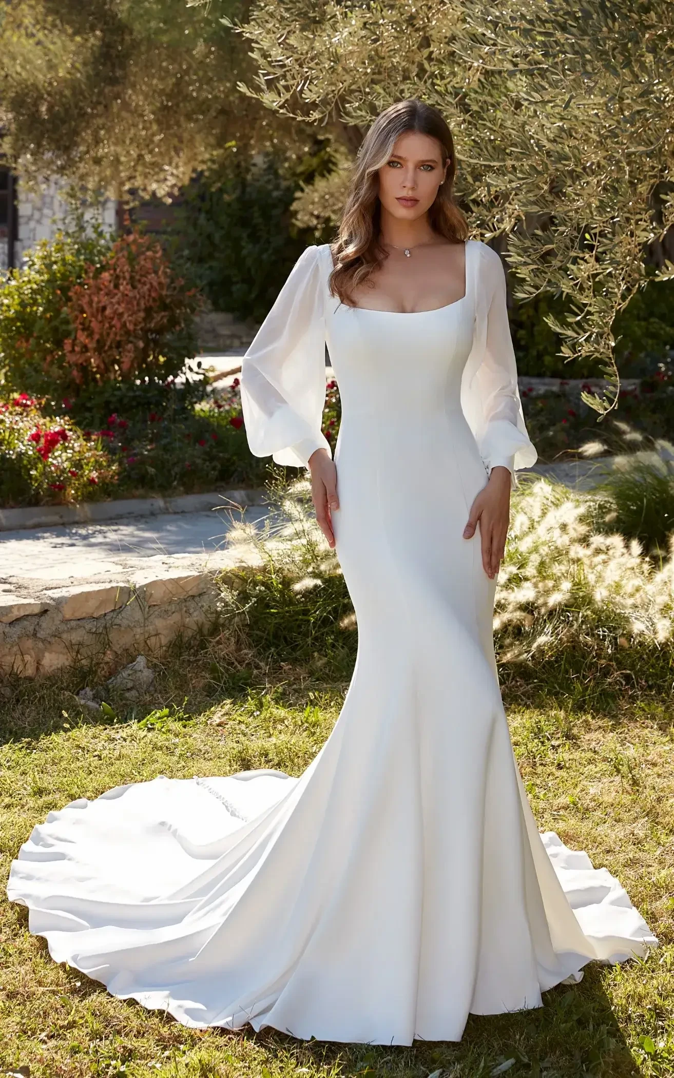 A woman in a white wedding dress with long, flowing sleeves stands in a garden with greenery and flowers in the background.
