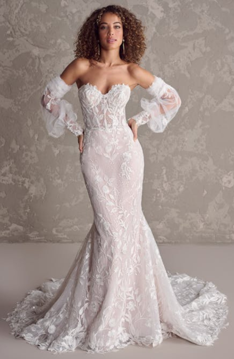 Woman in a strapless lace wedding gown with sheer, puffed sleeves, posing against a textured gray backdrop. The dress features floral patterns and a mermaid silhouette, exuding elegance and sophistication.