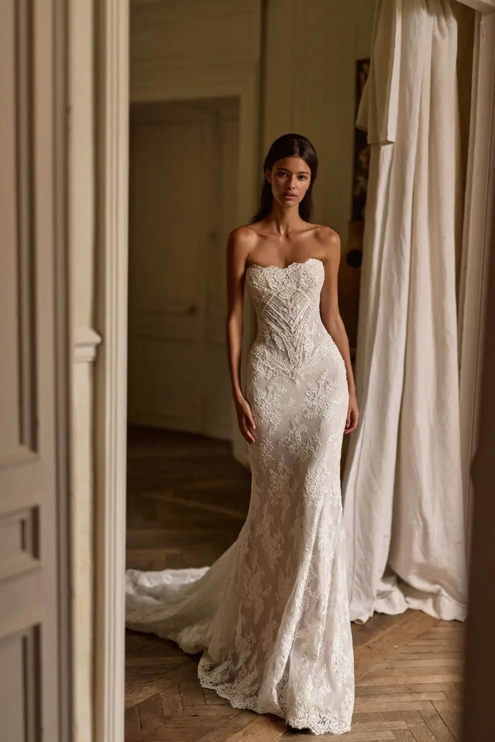 A model wearing a strapless, fitted lace wedding dress, standing in a room with natural light and elegant decor.