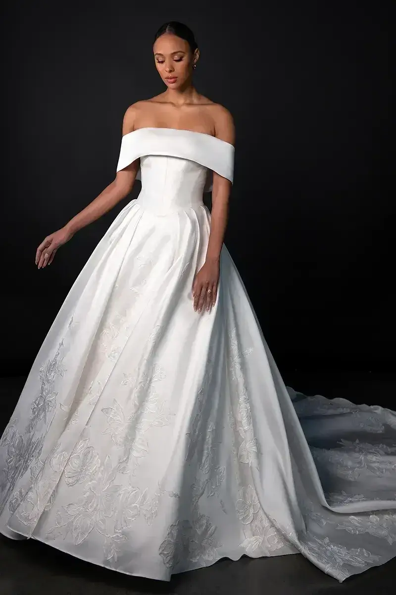 Model is wearing a princess-style ballgown with a straight-across neckline, off-the-shoulder straps, floral lace appliqués, hidden side pockets, and a long buttoned train.