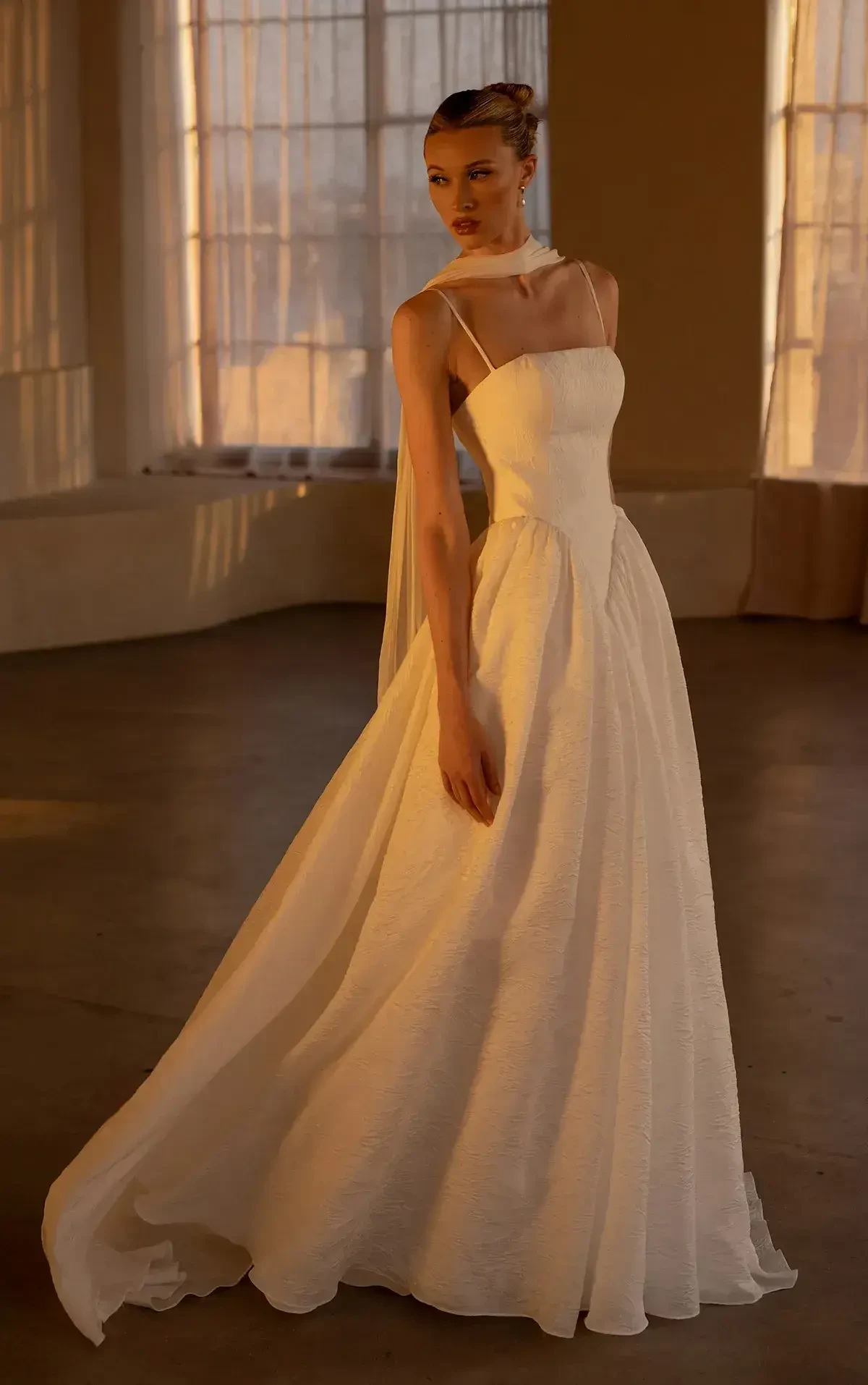 A model wearing a flowing white wedding dress with a fitted bodice and a long train, posed in an elegant indoor setting with soft lighting.
