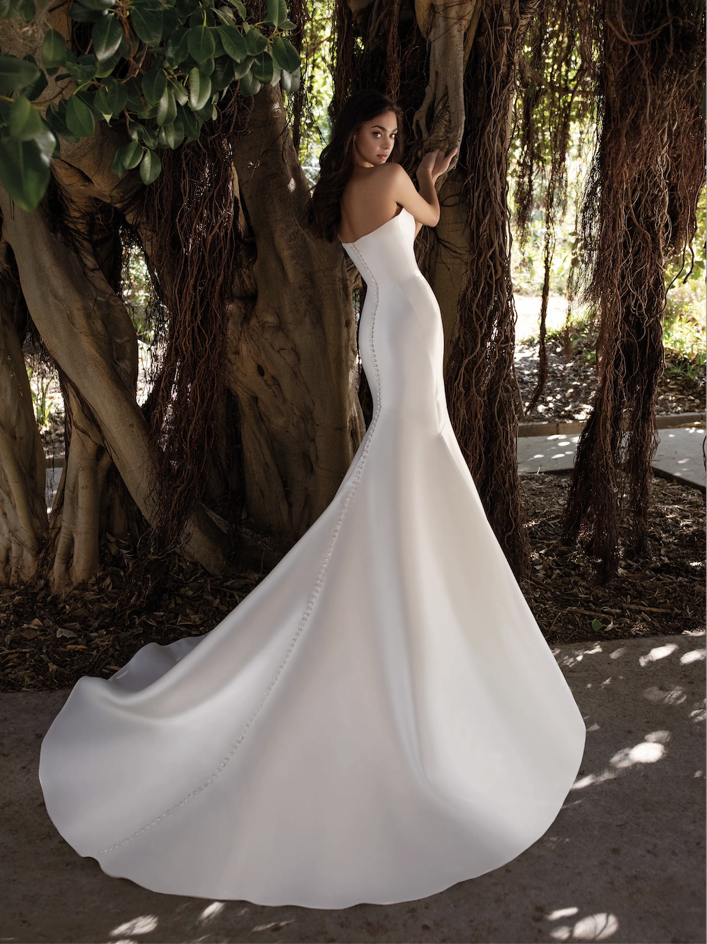 Model is wearing a strapless mikado trumpet gown with sculpted seams and elegant floor-length buttons trailing down the back.