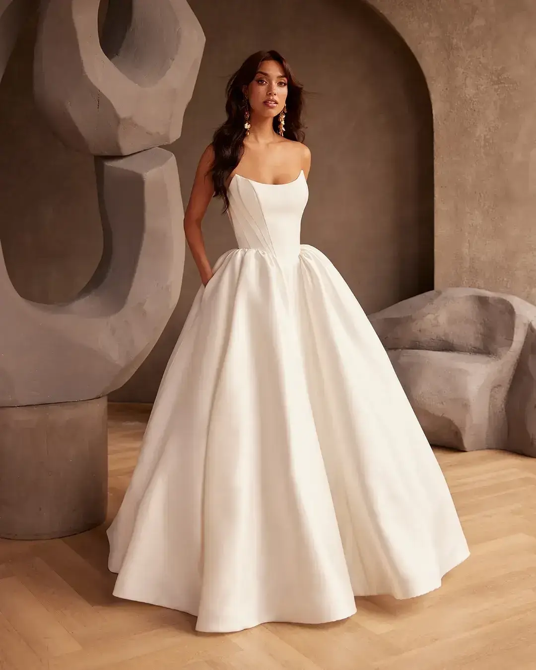 Model is wearing a strapless ballgown with a Mikado bodice, full gathered skirt with pockets, and an Italian tulle back featuring exposed boning and fabric-covered buttons.