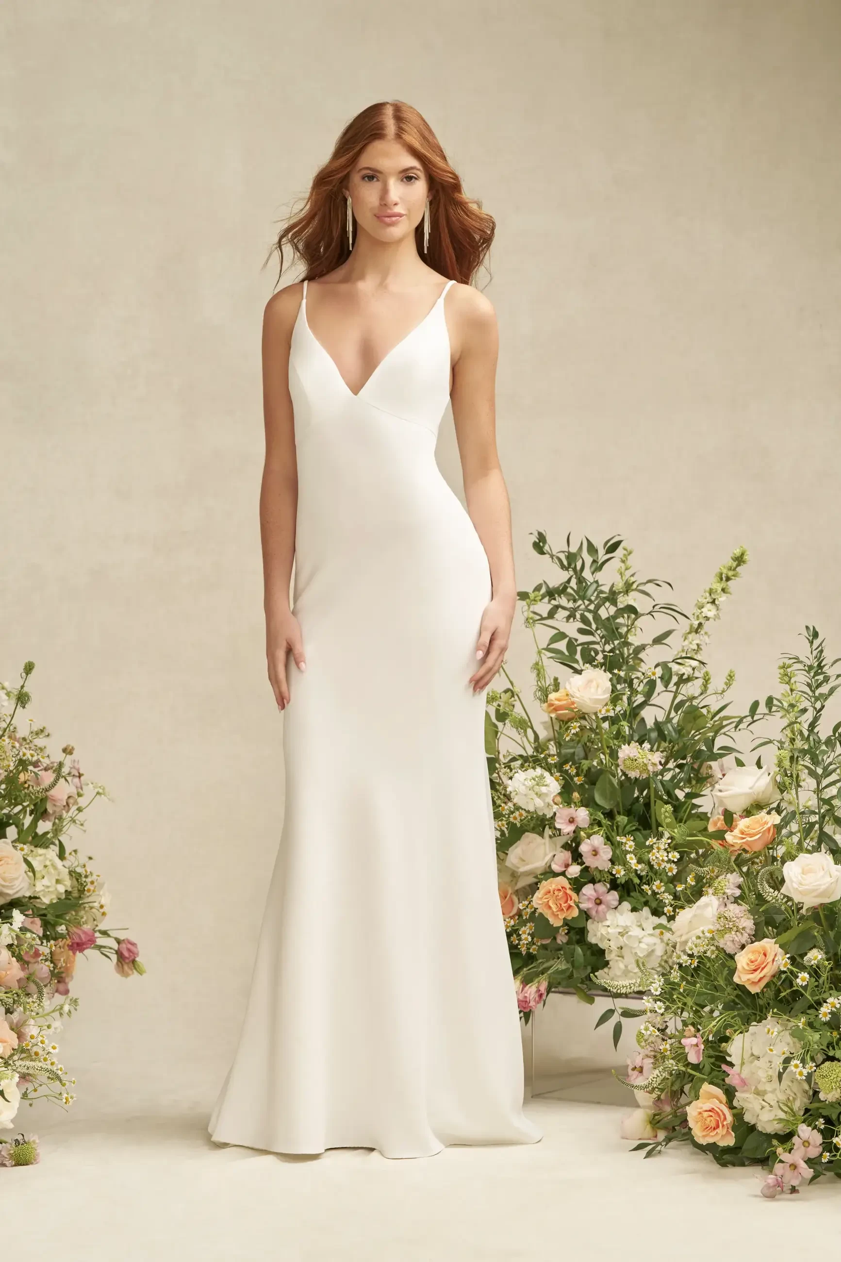 A model wearing a sleek, white evening gown stands in front of floral arrangements featuring various pastel flowers.