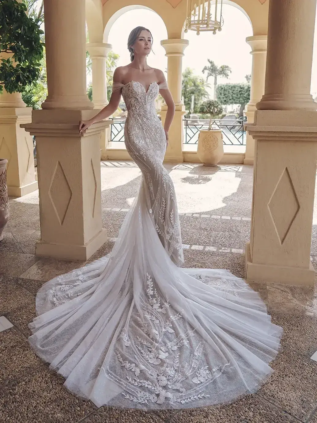 Model is wearing a mermaid gown with sequin-beaded floral lace, an illusion corset back, detachable off-the-shoulder tulle straps, and an elongated train.