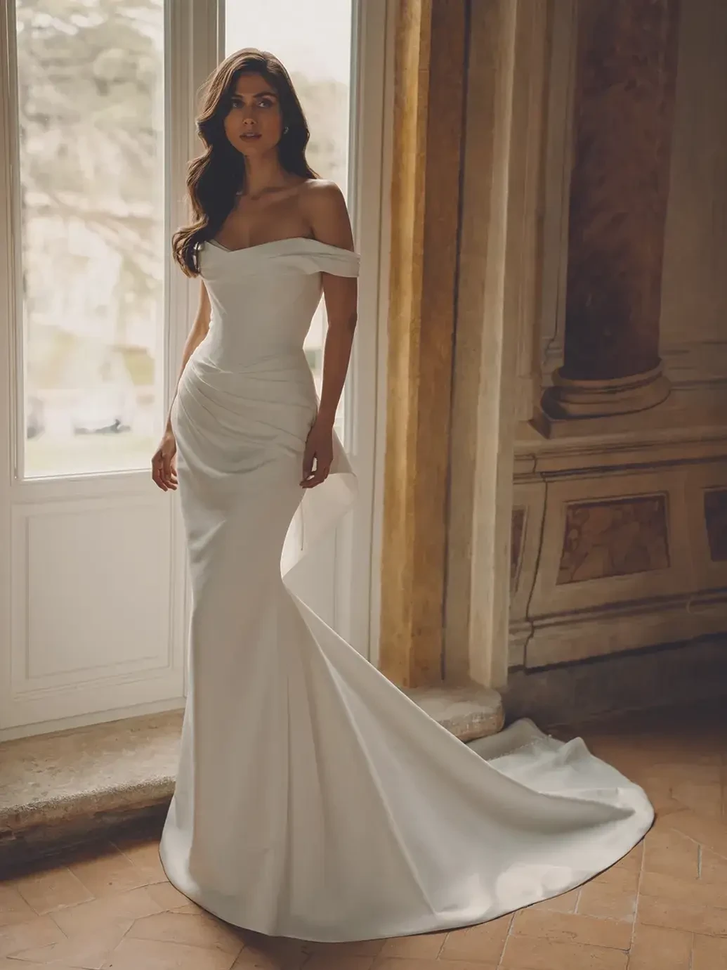 A model wearing a stunning off-the-shoulder white wedding gown stands elegantly in front of a large window, showcasing the dress's fitted silhouette and flowing train. The setting features classic architecture with warm tones and soft natural light.