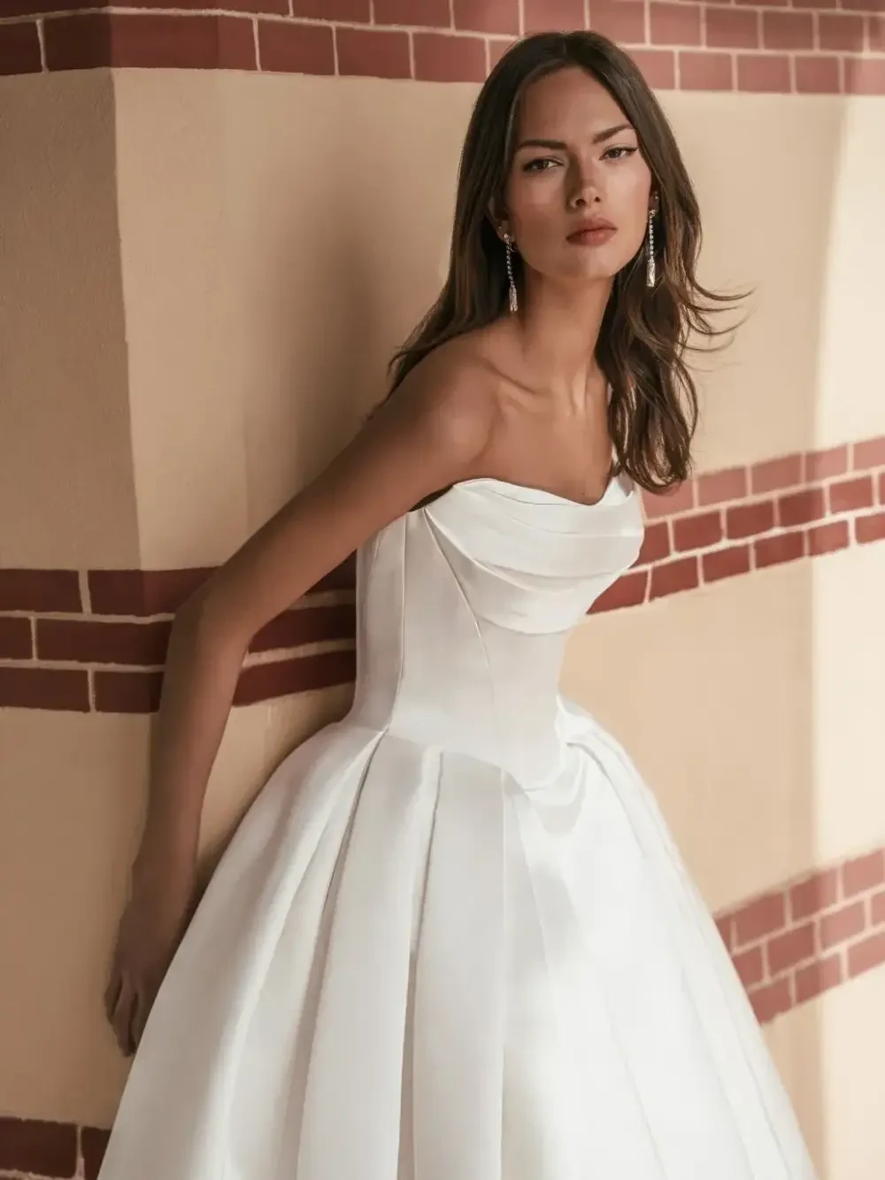 A woman in a strapless white gown poses elegantly against a textured wall.